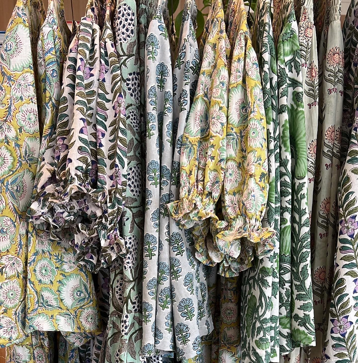 A close-up of multiple patterned bohemian-style blouse or dress panels hanging on a rack. The fabrics feature tropical floral and leaf prints in green, yellow, purple, and blue on cream backgrounds.